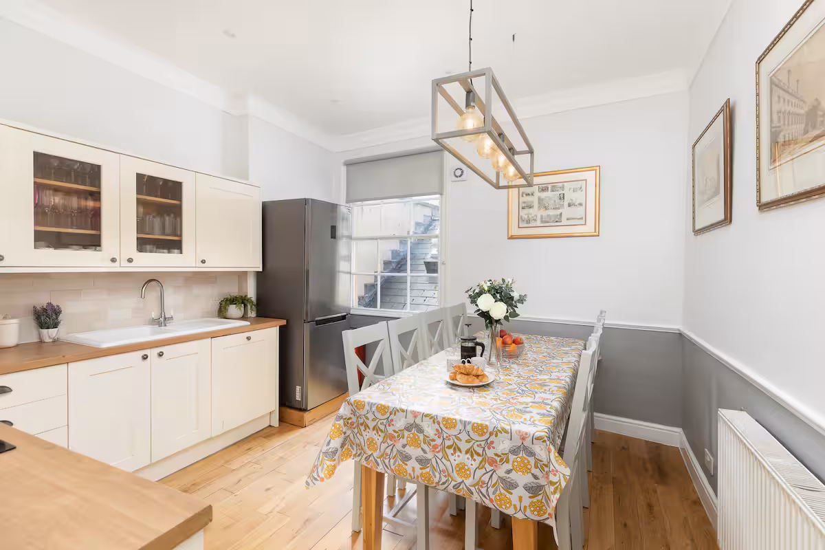 Open-plan kitchen-dining with cream cabinets, sunken extension and an oak dining table dressed in a yellow floral oilcloth under a lantern pendant