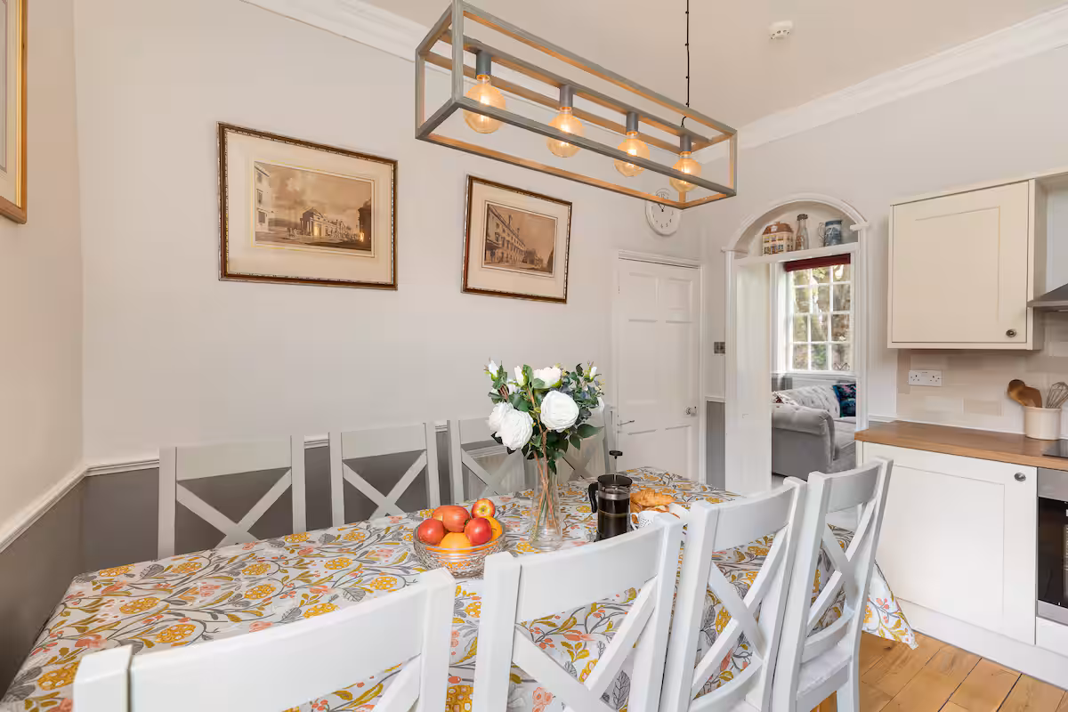 Dining table set for six with white cross-back chairs, oilcloth tablecloth, framed Bath prints on the wall and an arched doorway through to the lounge