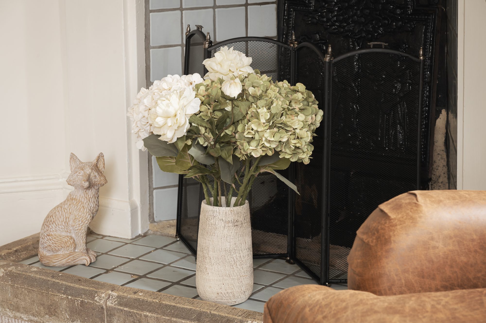 Close-up of white peonies and dried hydrangeas in a stone vase beside a cast-iron fireplace