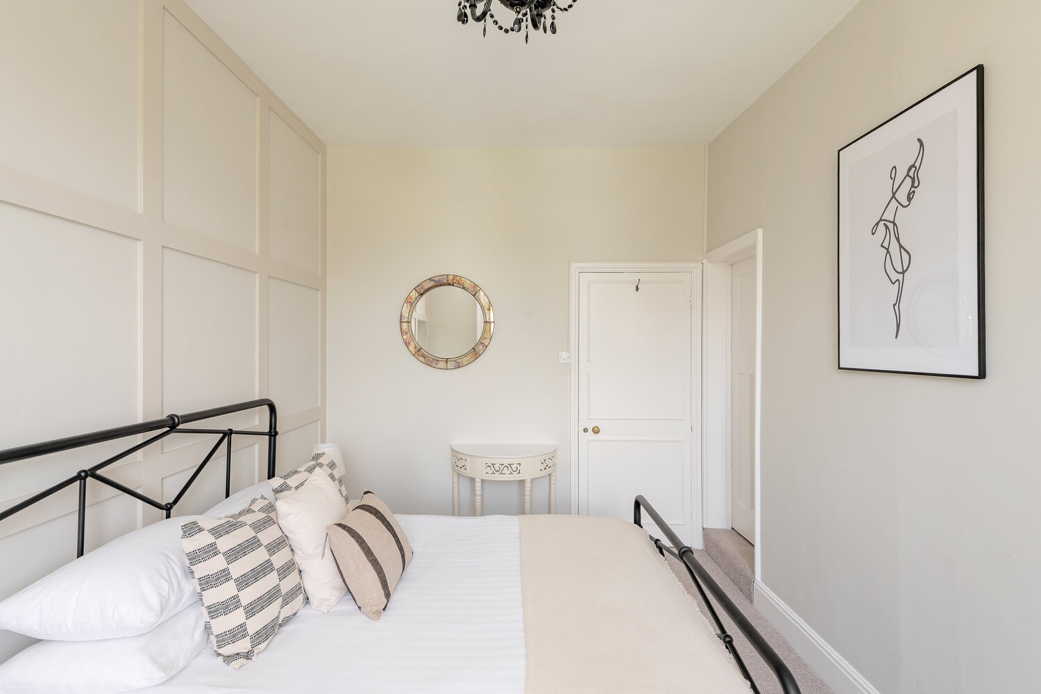 Cream panelled bedroom with black metal bedframe, striped pillows, black chandelier and a round mirror above a small white console table.