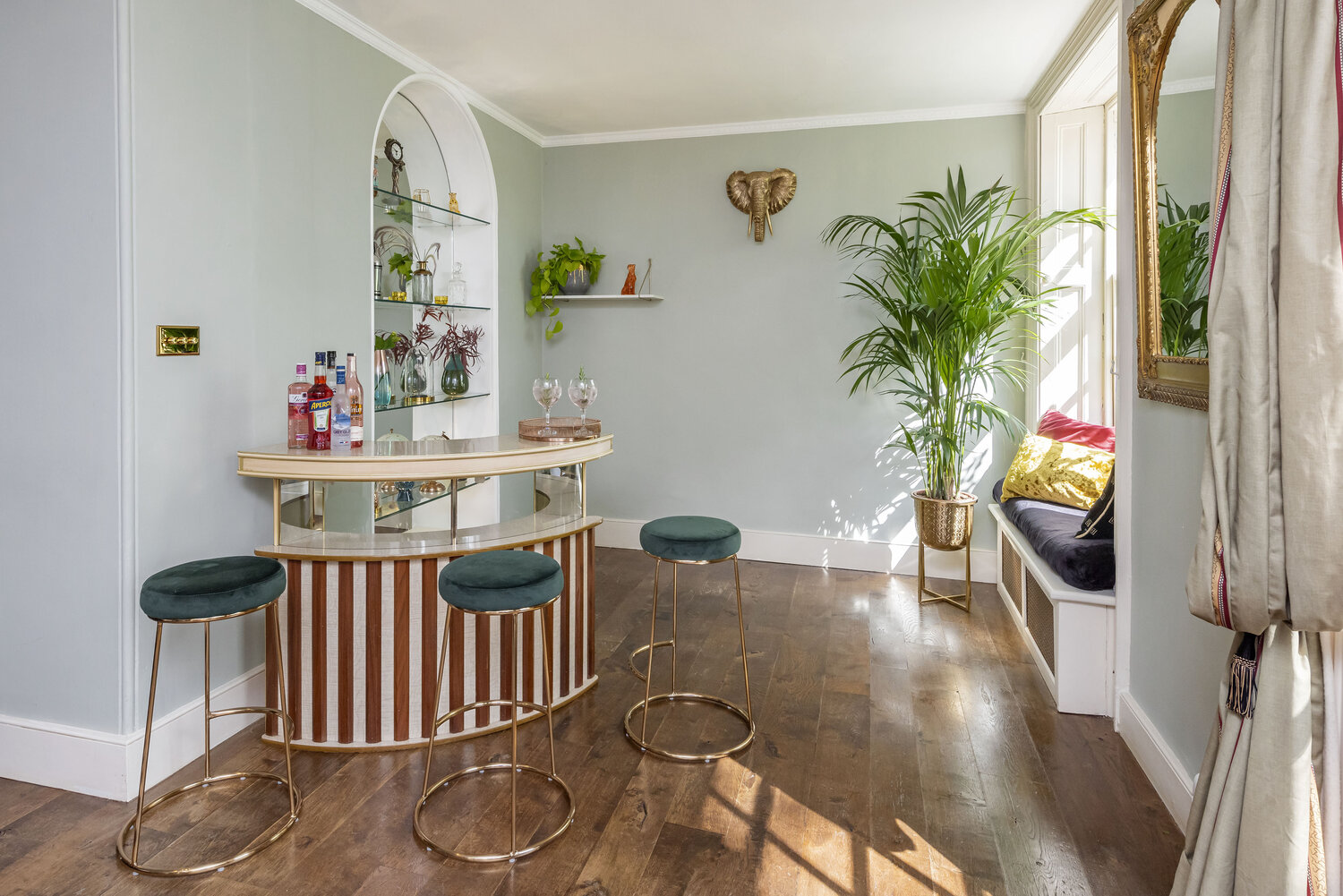 Sage green bar nook with a curved striped wood bar, teal velvet barstools on brass legs, drinks bottles and a tall potted palm by the window.