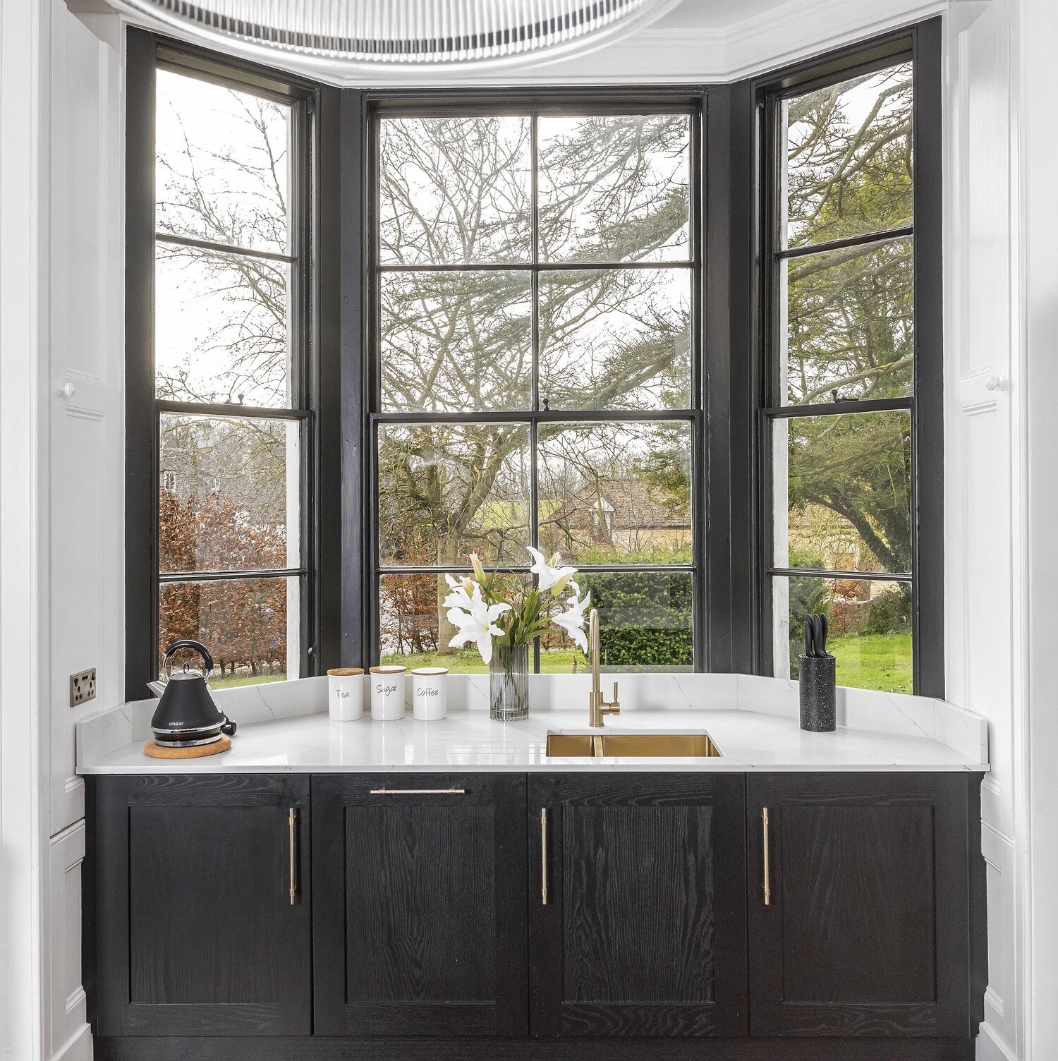 Bay window prep kitchen with black sash windows over the garden, white worktop, brass tap, lilies in a glass vase and labelled tea, sugar, coffee tins.