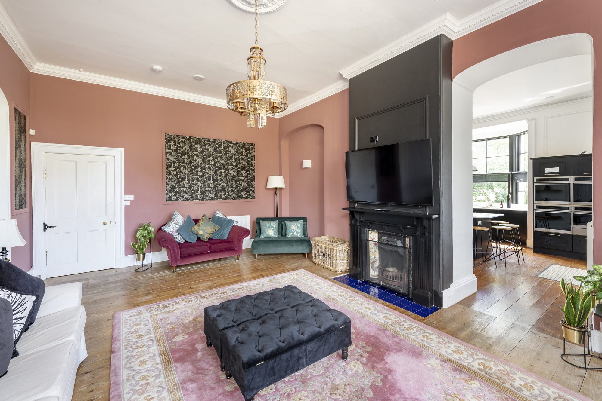 Dusty pink drawing room with gold chandelier, black tufted ottoman on a faded pink rug, plum and teal velvet sofas and a black fireplace under a wall-mounted TV.