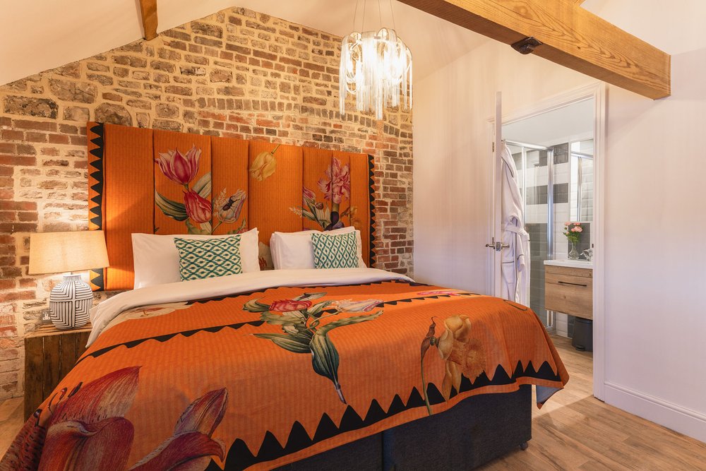 Brick-walled bedroom with orange tulip-pattern headboard, vaulted oak beams and ensuite door