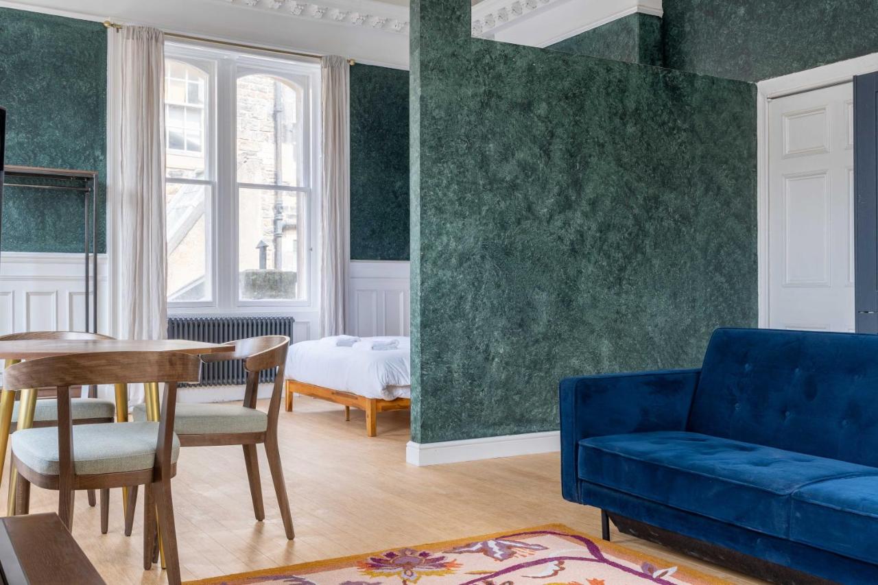 Open-plan green-walled studio with double bed in bay window, dining table and blue velvet sofa