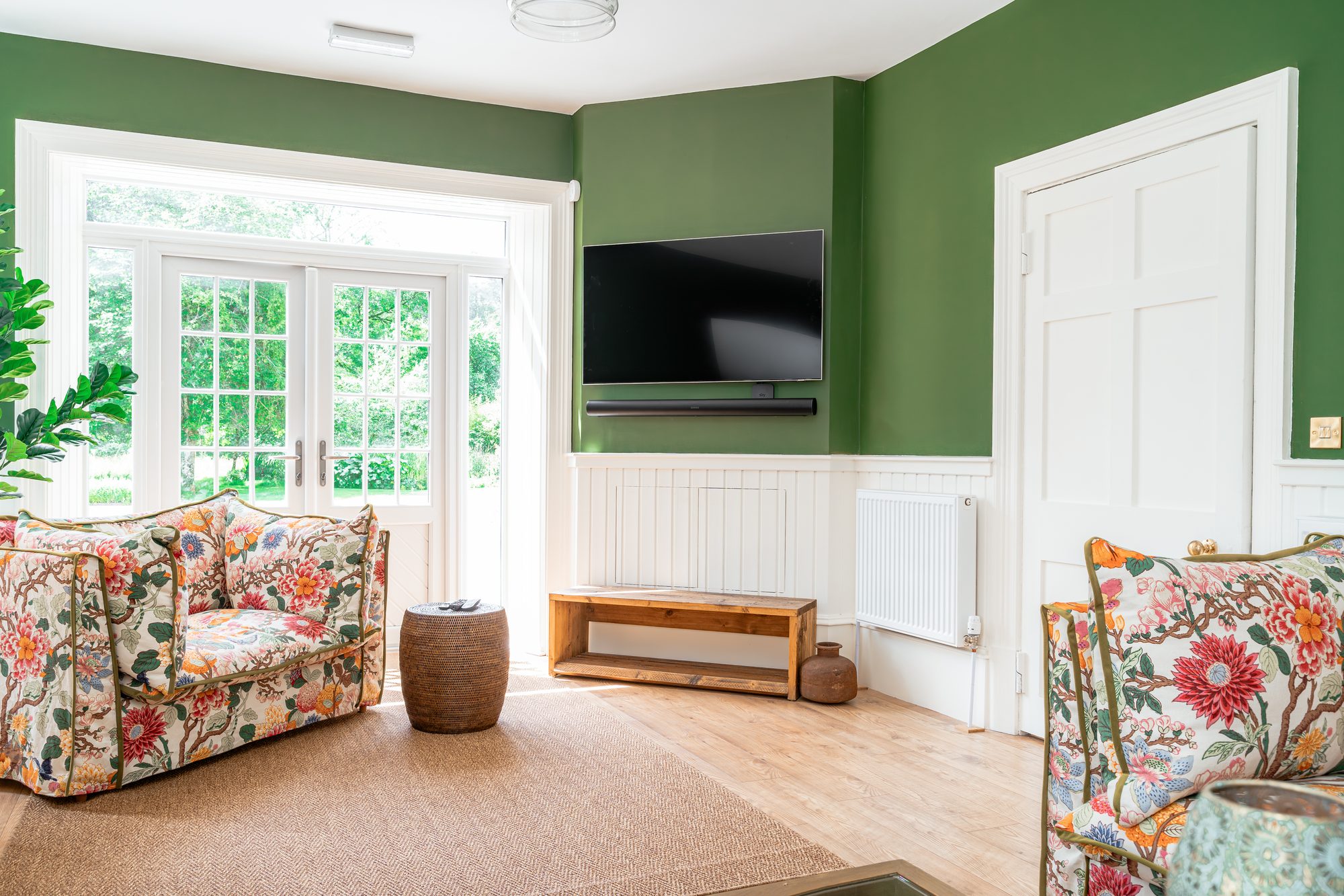 Green TV snug with floral patterned sofas, white panelling, wall-mounted television and French doors opening onto the garden