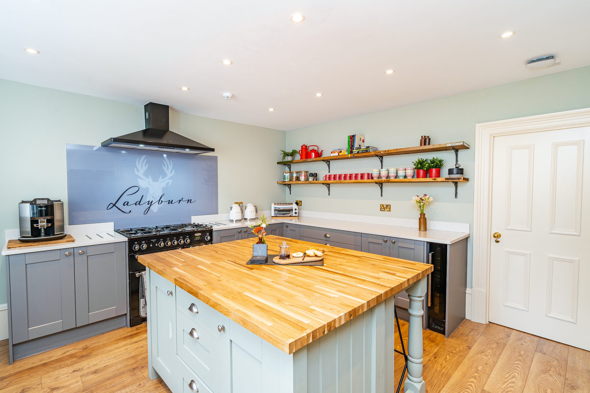 Sage green country kitchen with central oak butcher block island, range cooker under a Ladyburn stag splashback and open shelving with red kettles
