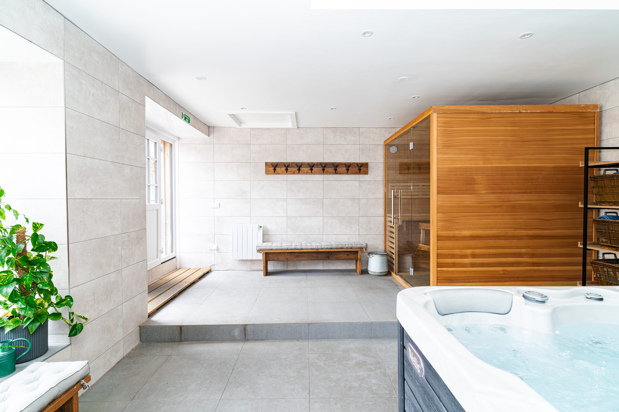 Spa room with cedar sauna, deep blue hot tub in the corner, tiled walls, antler coat hooks above a wooden bench and a leafy houseplant