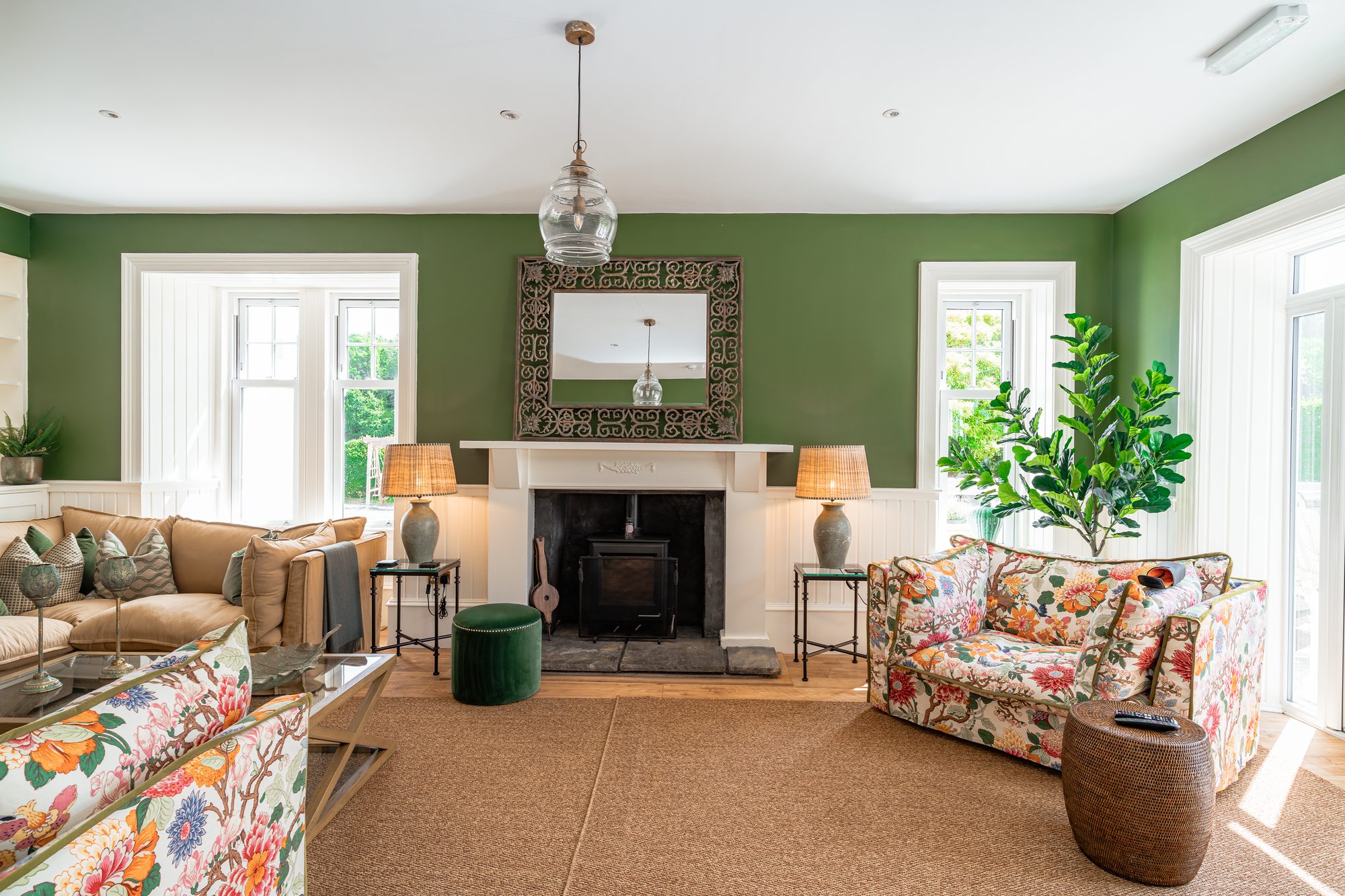 Green sitting room with floral sofas around a wood-burning stove, fiddle leaf fig, ornate Indian-style mirror above the mantel and bright French doors