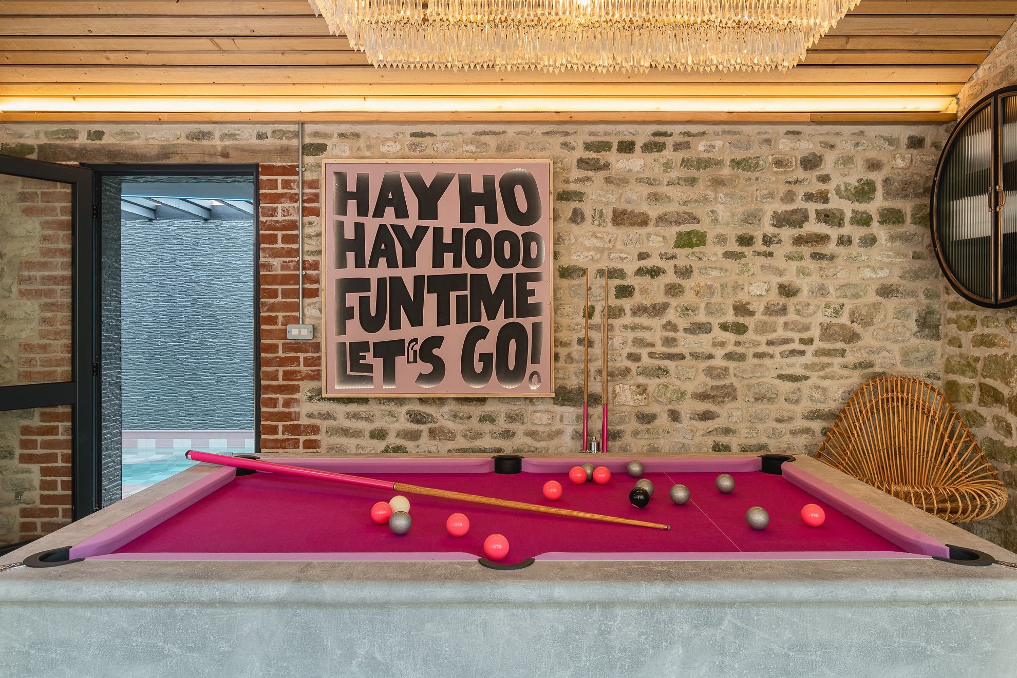 Pink-felt pool table at Country House Cool under a HAYHO HAYHOOD FUNTIME LET'S GO mural, with stone walls and a hanging chandelier