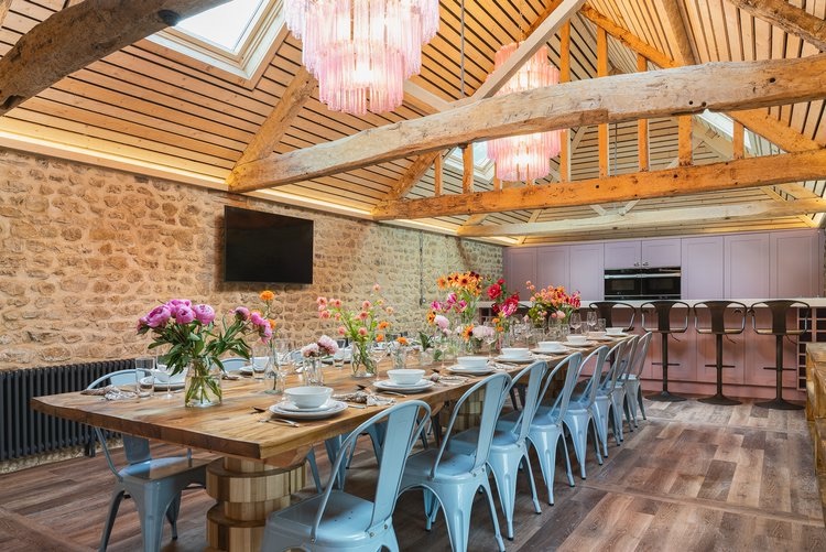 Long timber dining table set for sixteen with peonies and dahlias, blue Tolix chairs, pink glass chandeliers and pink-shaker kitchen beyond