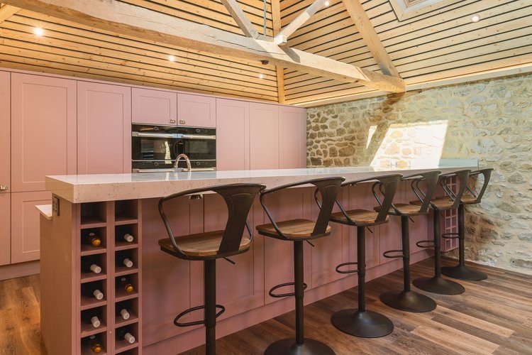 Dusty-pink shaker kitchen island with five metal bar stools, integrated wine rack and exposed timber beams above