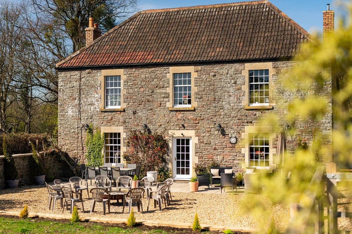Stone Georgian farmhouse front on a sunny day with tall sash windows and an outdoor dining table set on the gravel