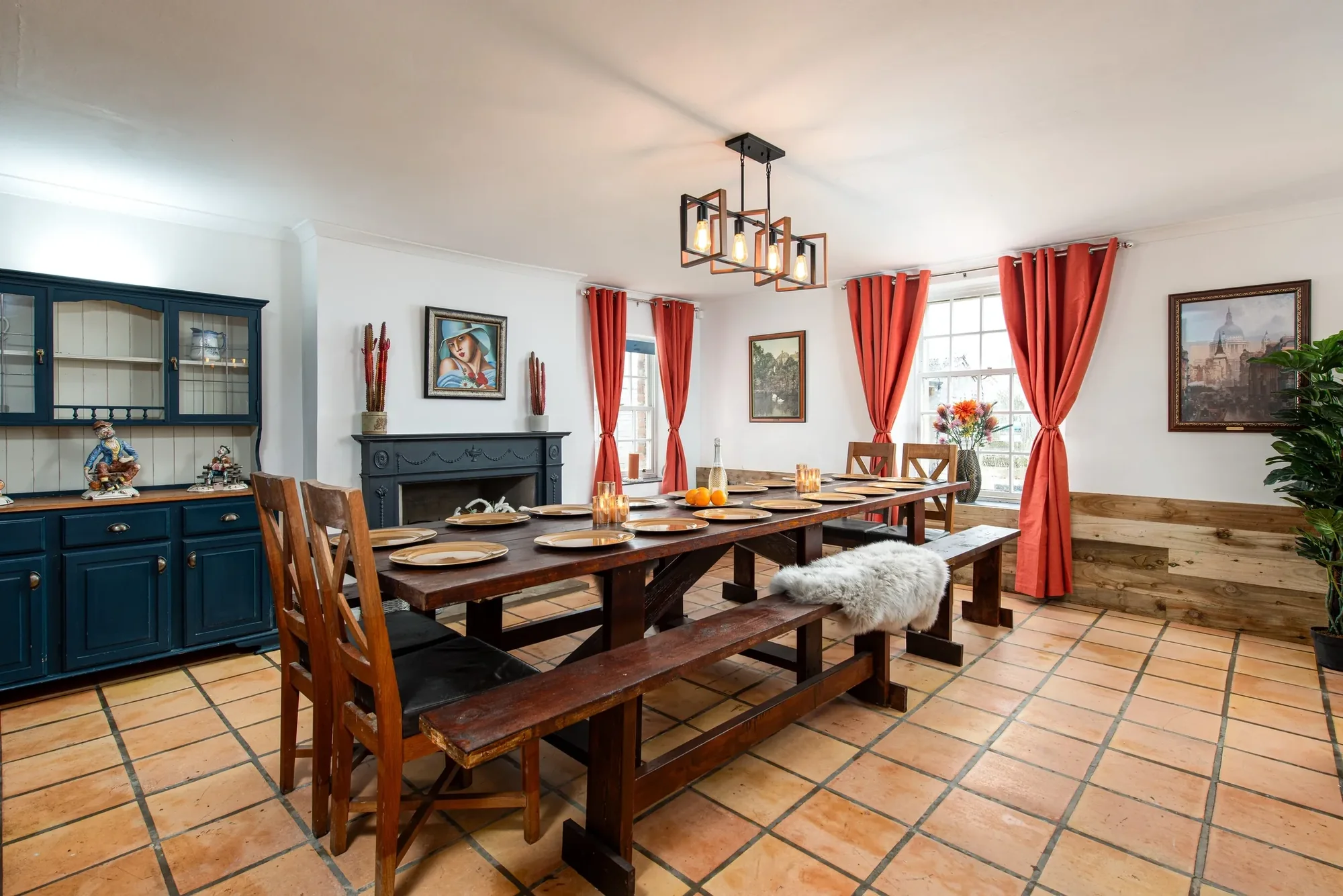 Interior dining room with a long farmhouse table, blue dresser, red curtains and a framed portrait above the fireplace