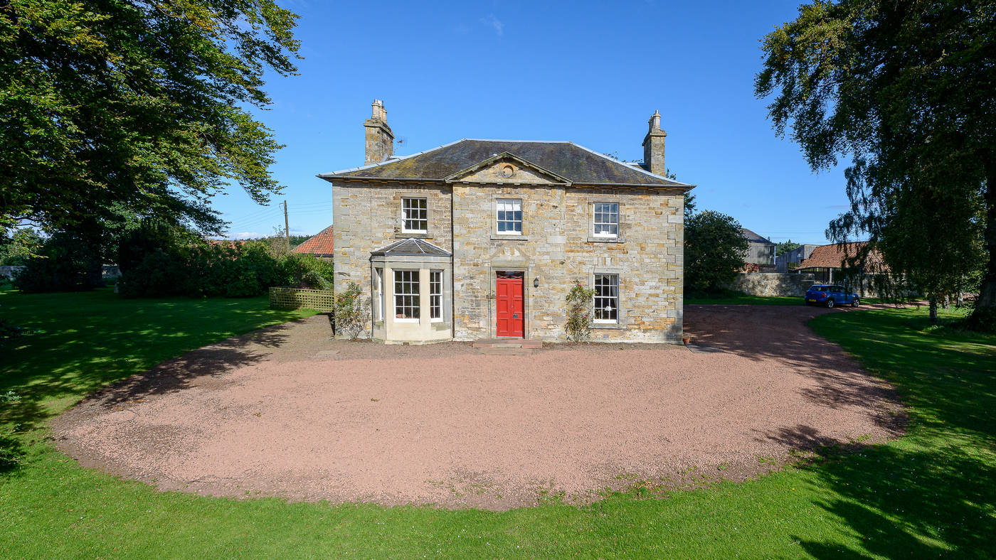 Symmetrical stone Georgian farmhouse with red front door, lawns and a sweeping gravel drive