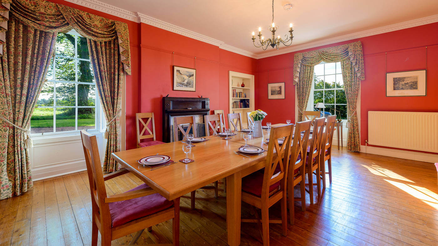 Red dining room with long oak table for ten, swag curtains and tall sash windows over fields