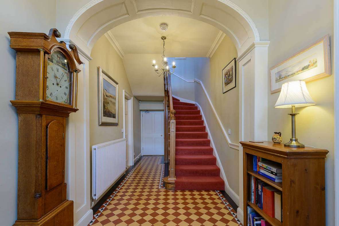 Hall with tiled floor, grandfather clock and red carpeted staircase under arched cornicing