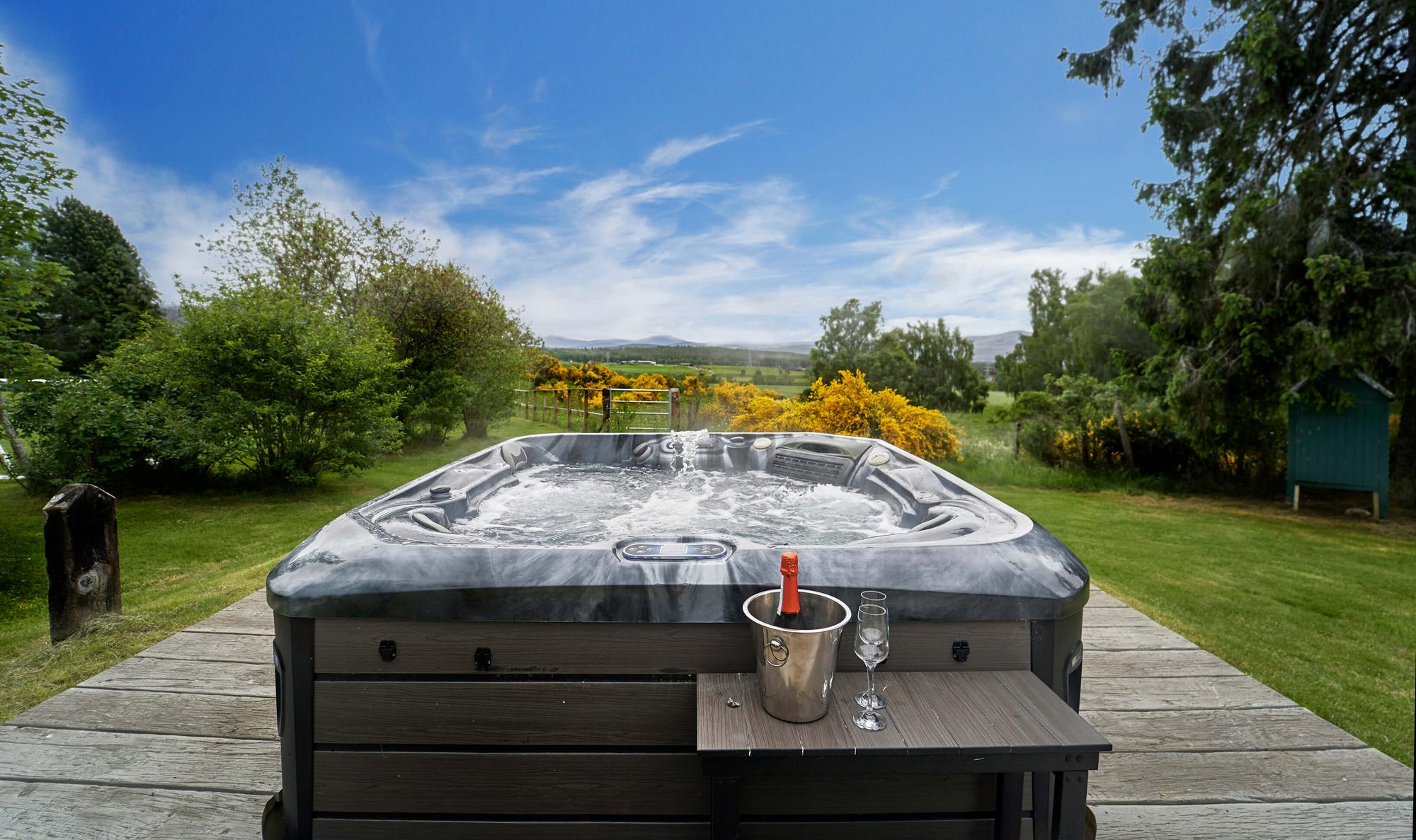 Hot tub on a deck with prosecco and glasses, framed by gorse, hills and a wide blue sky
