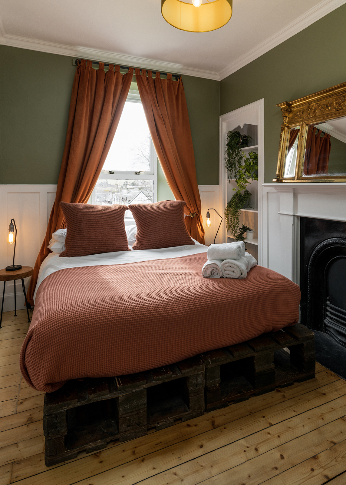 Bedroom in olive green and pink with rust velvet curtains, gold mantel mirror, original cast iron fireplace and a pallet bed