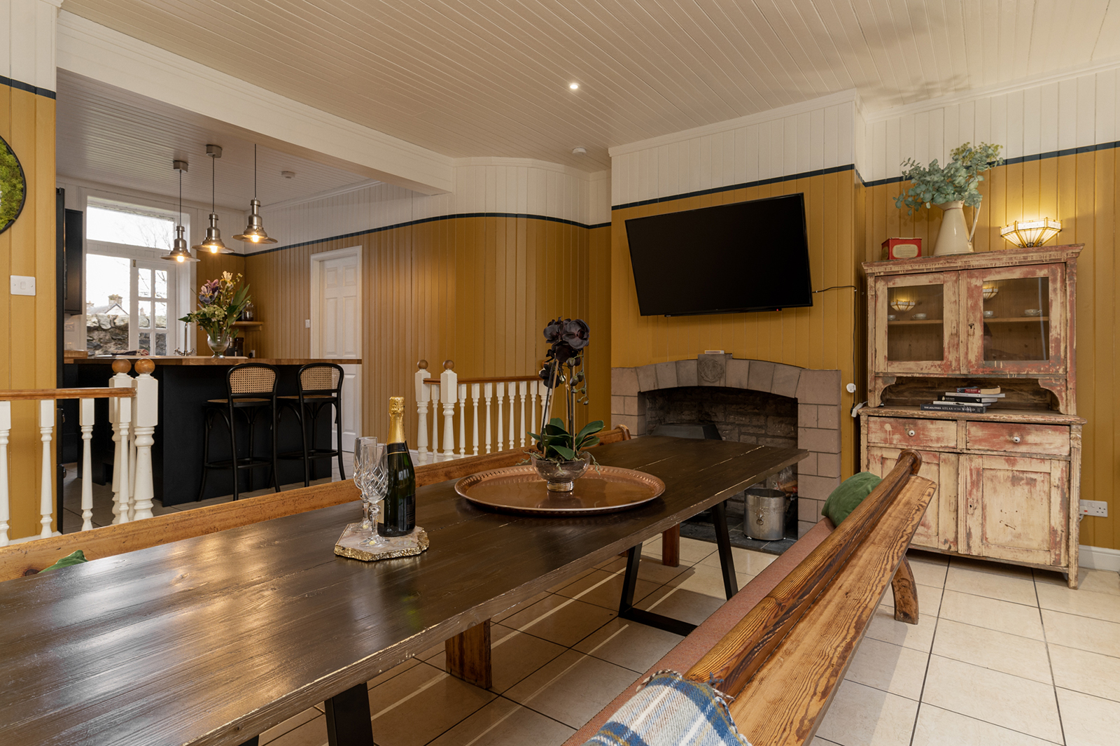 Mustard yellow panelled dining room with long farmhouse table, bench seating, mounted TV and a vintage painted dresser