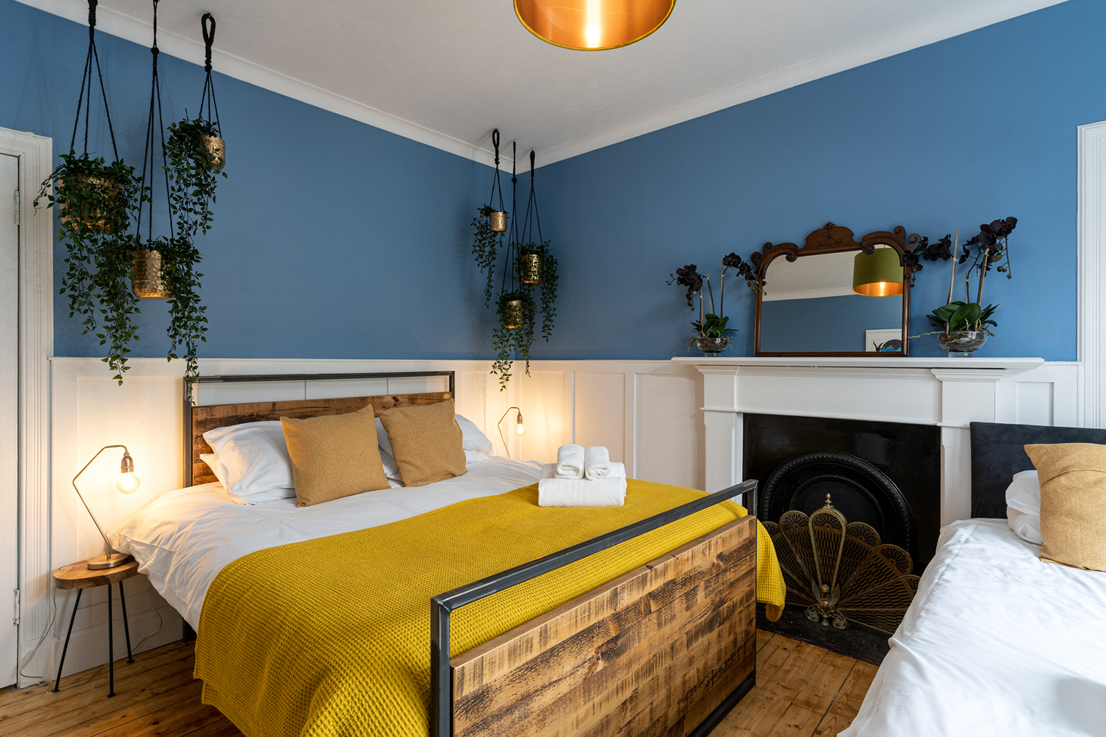 Twin bedroom with deep blue walls, hanging brass plant pots, panelled wainscoting, ornate mirror and a reclaimed wood bed frame