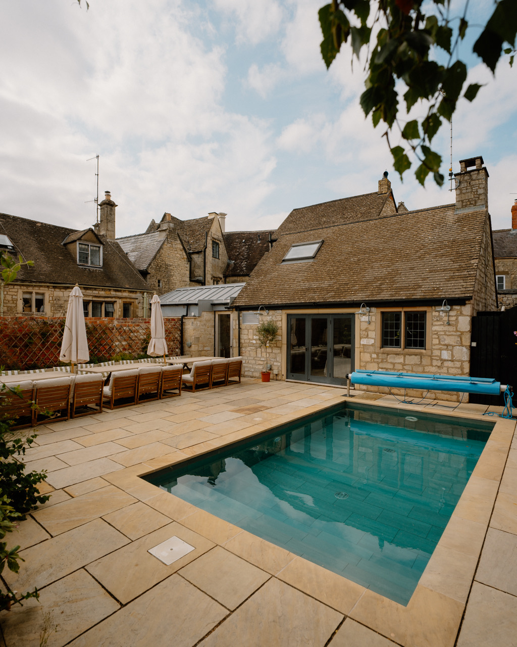 Heated outdoor pool on a pale stone terrace with Cotswold-stone cottages and trees behind