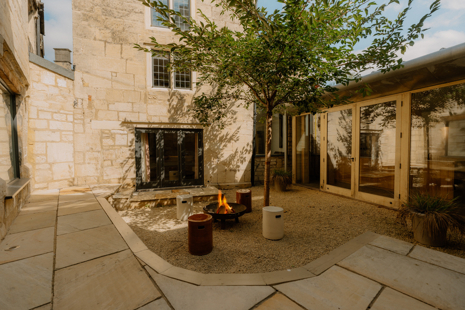 Sunlit gravel courtyard with a tree, stone seating ledge and a lit fire pit framed by honey-stone walls and glass doors