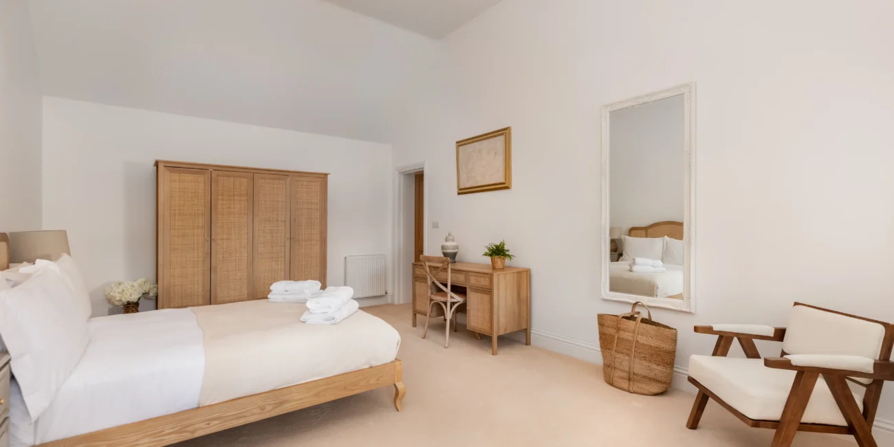 White double bedroom with rattan wardrobe, gold-framed mirror and towel stack on the bed