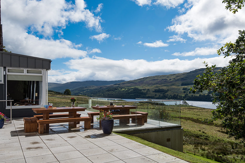 Outdoor terrace with two long picnic tables, glass balustrade and uninterrupted view down the loch