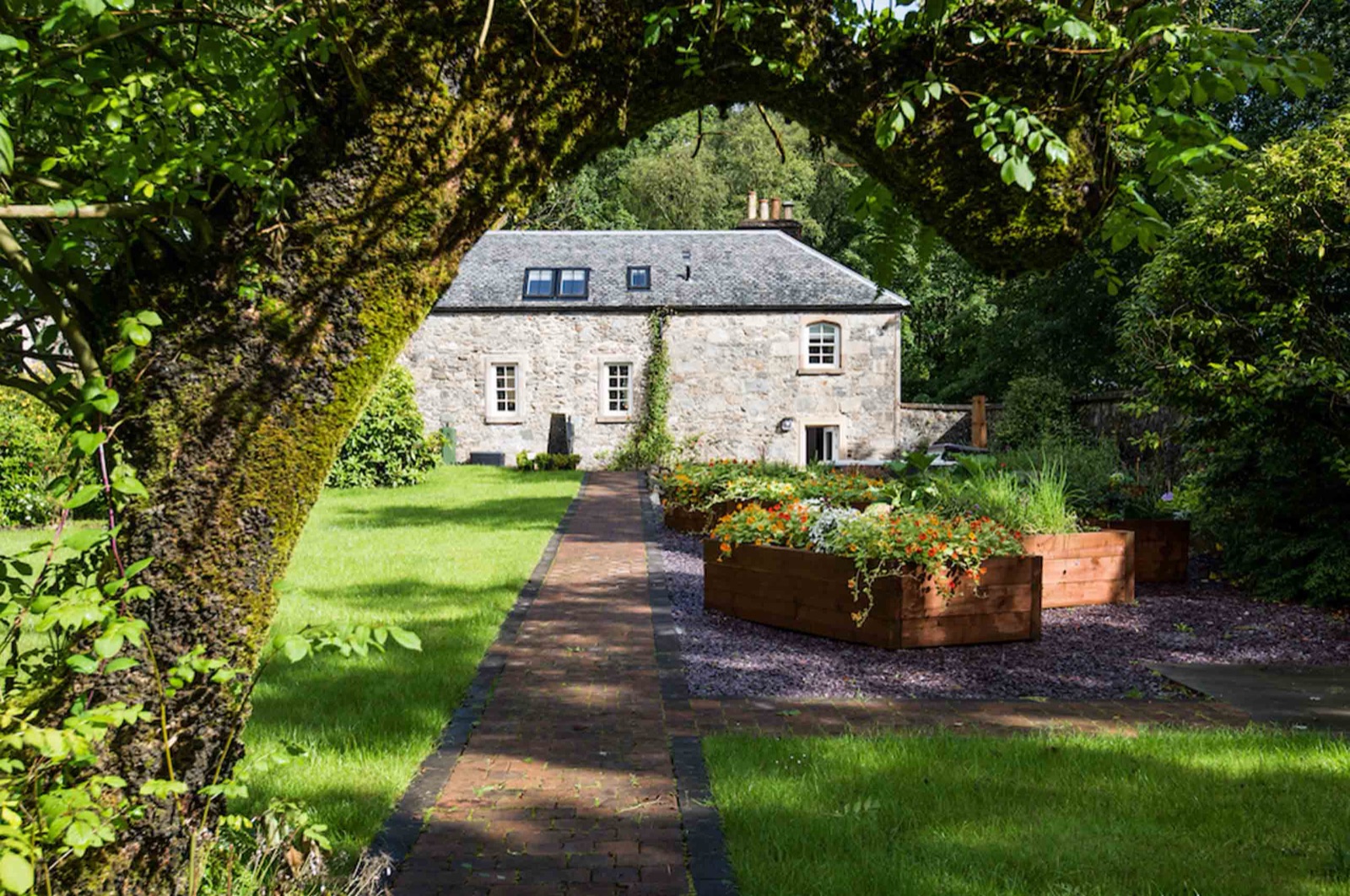 Stone house framed by a mossy tree arch with a brick path and raised flower beds