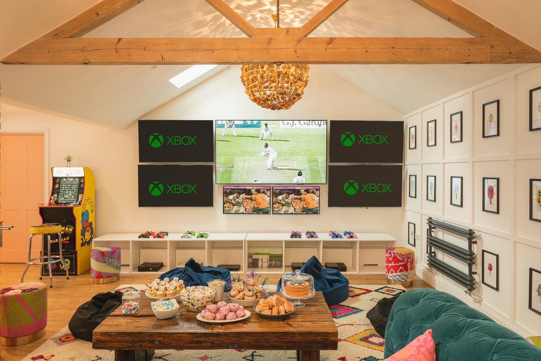 Cinema-and-games loft under vaulted beams with three Xbox screens, arcade cabinet and a dessert spread on a low wooden table