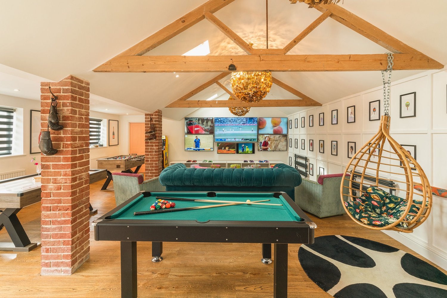 Games room with green-felt pool table, hanging rattan chair, foosball, wall of TVs and brass-bead chandelier under vaulted beams