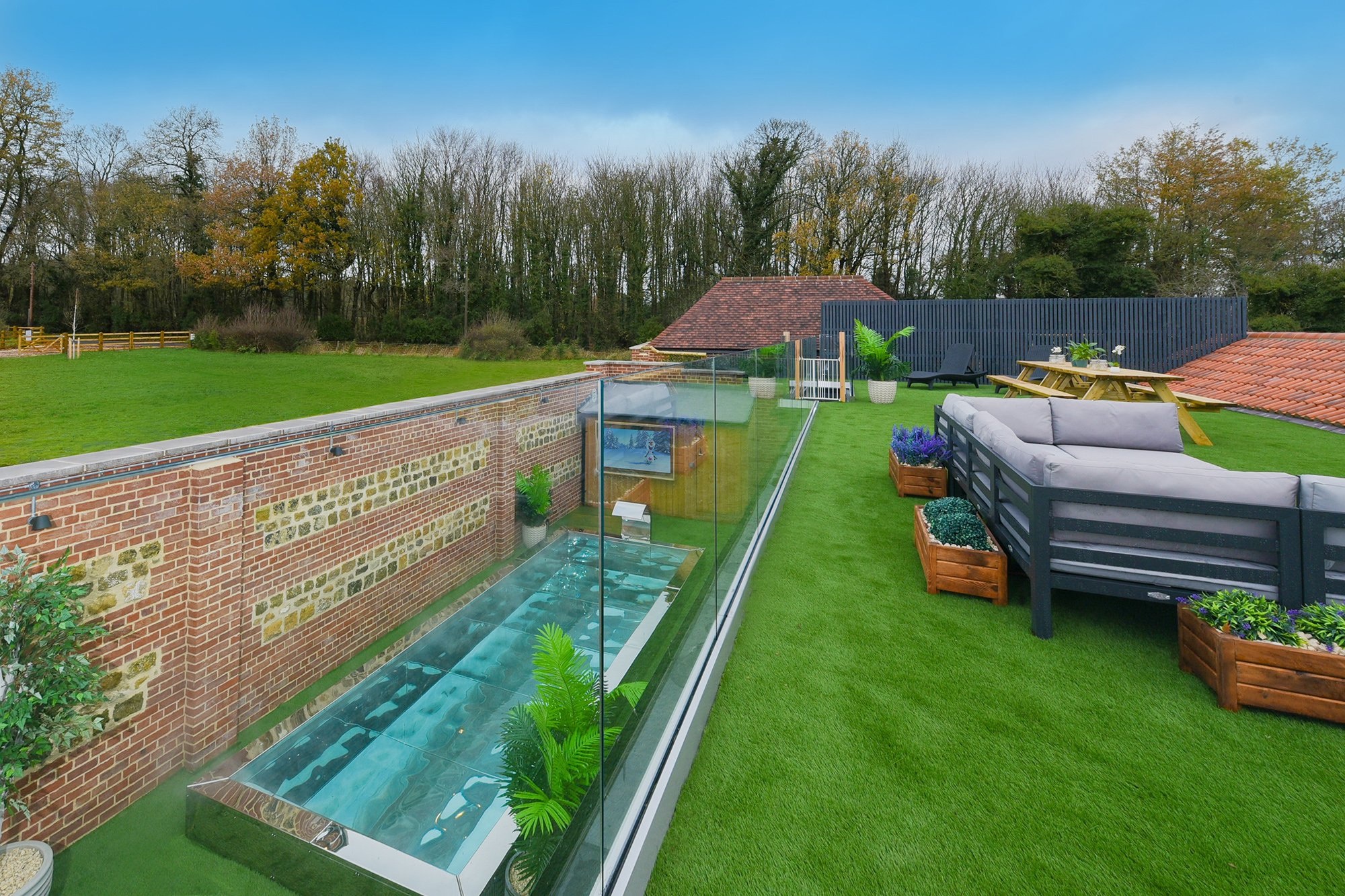 Daytime view of the outdoor pool with bubble jets, glass-walled, with garden sofa lounge and planters on the lawn alongside