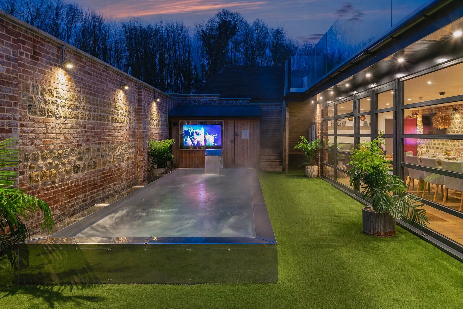 Outdoor heated pool at dusk with a big outdoor cinema screen on the exposed stone-and-brick wall, artificial lawn and the glass-walled house lit up alongside