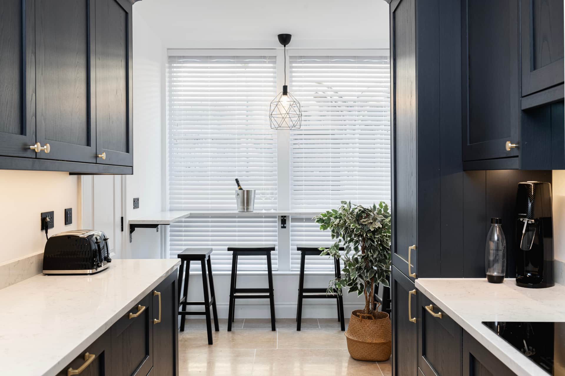 Navy shaker galley kitchen with brass handles, white quartz worktops and a breakfast bar with three black stools