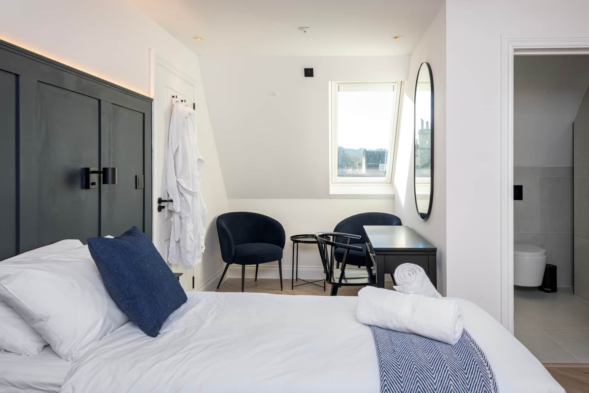 Top-floor bedroom with black panelled headboard, oval mirror and a dormer window onto Bath rooftops