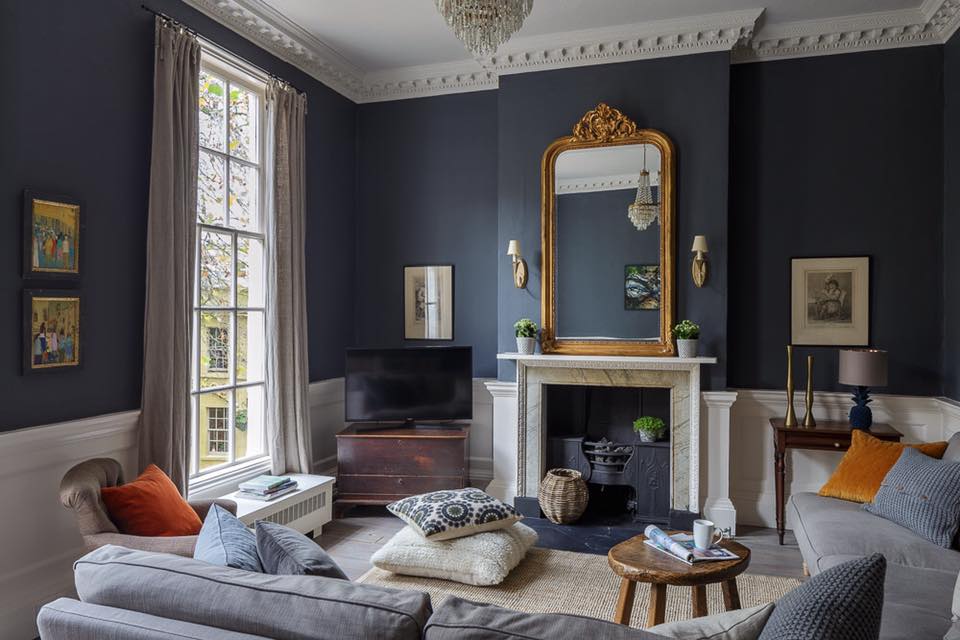 Navy lounge with white wainscot, marble fireplace under a gilded mirror, two grey sofas and a chandelier