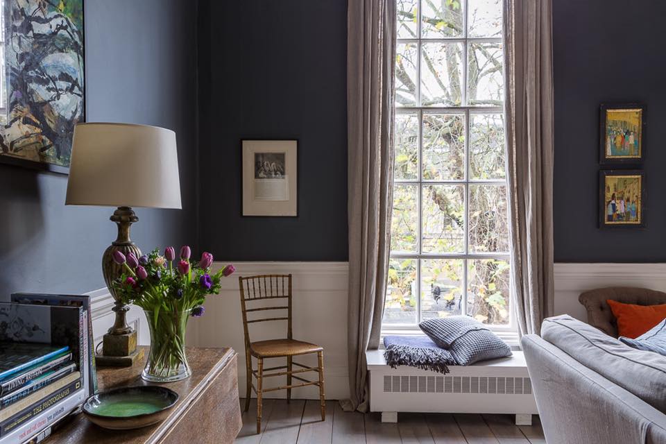Sitting room corner with brass lamp, tulips on a console table and a cushioned window seat by a tall sash window
