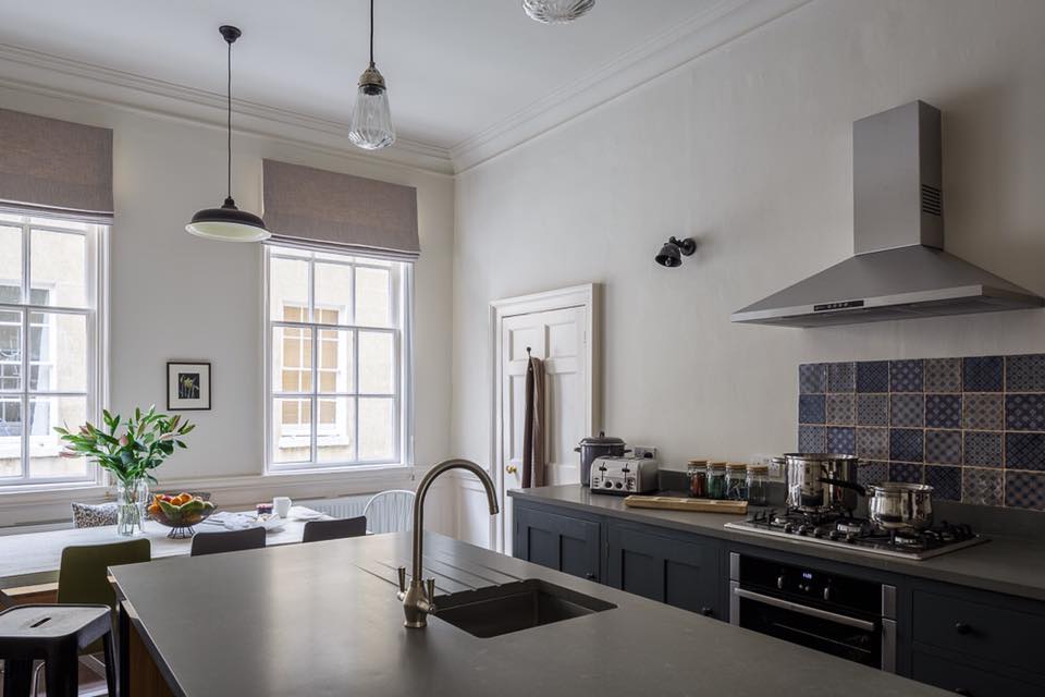 Kitchen island with gas hob, blue-tiled splashback and dining table set up by tall sash windows