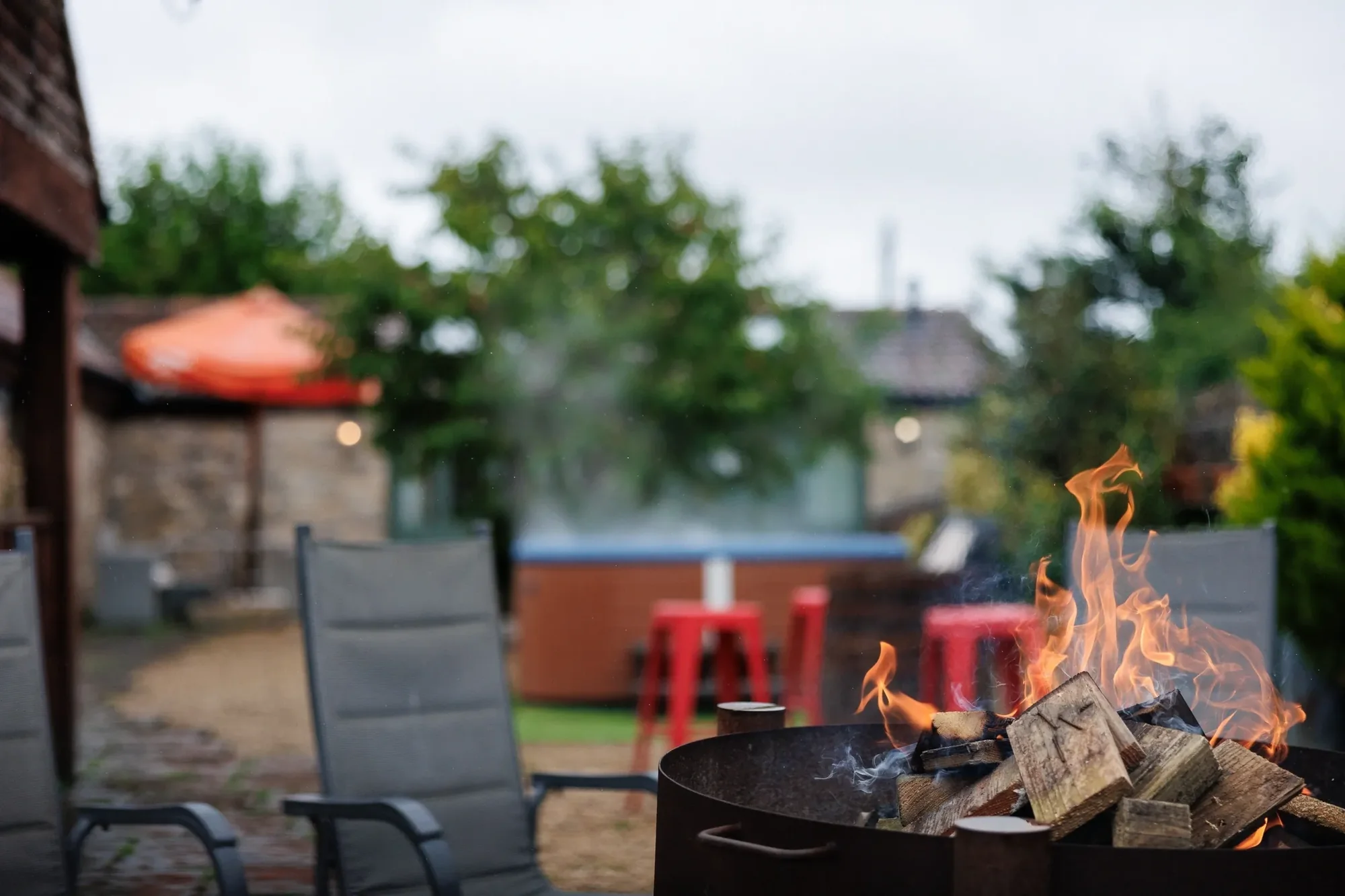 Open log fire-pit in the foreground with hot tub and red bar stools blurred behind in the courtyard