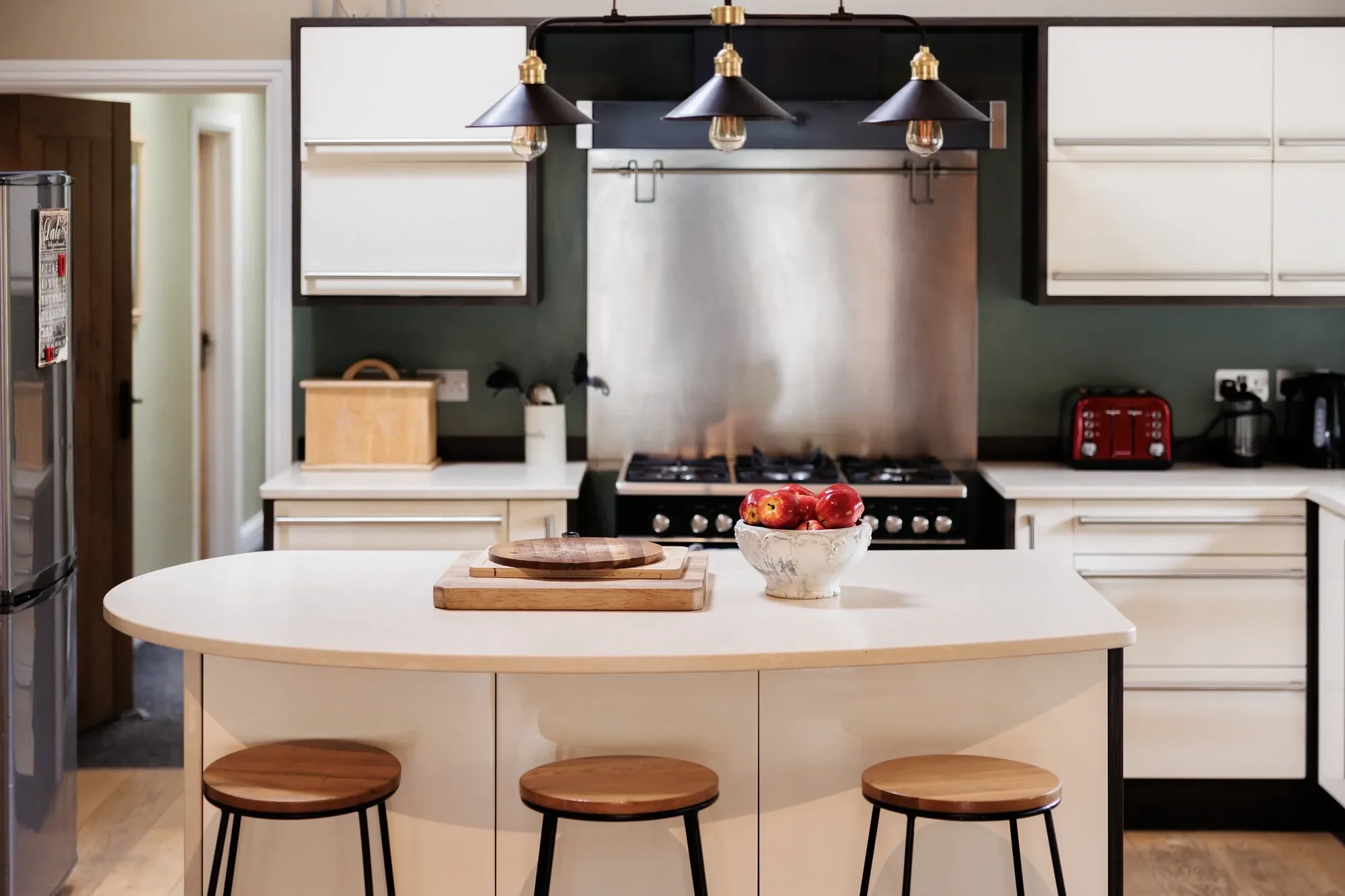 Kitchen with rounded cream island, wooden stools and triple black pendant lights over a range cooker