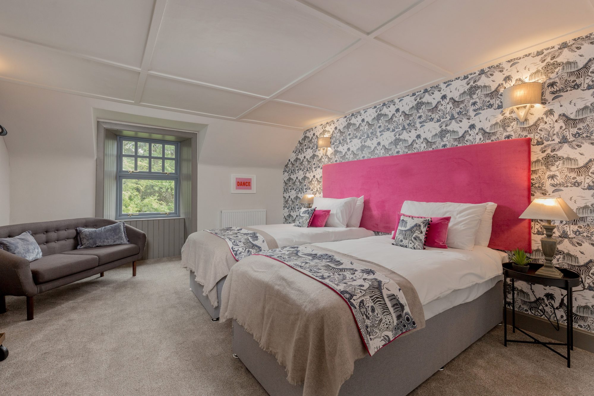 Twin-bed room with bright pink velvet headboard against zebra-and-jungle wallpaper, plus a grey sofa