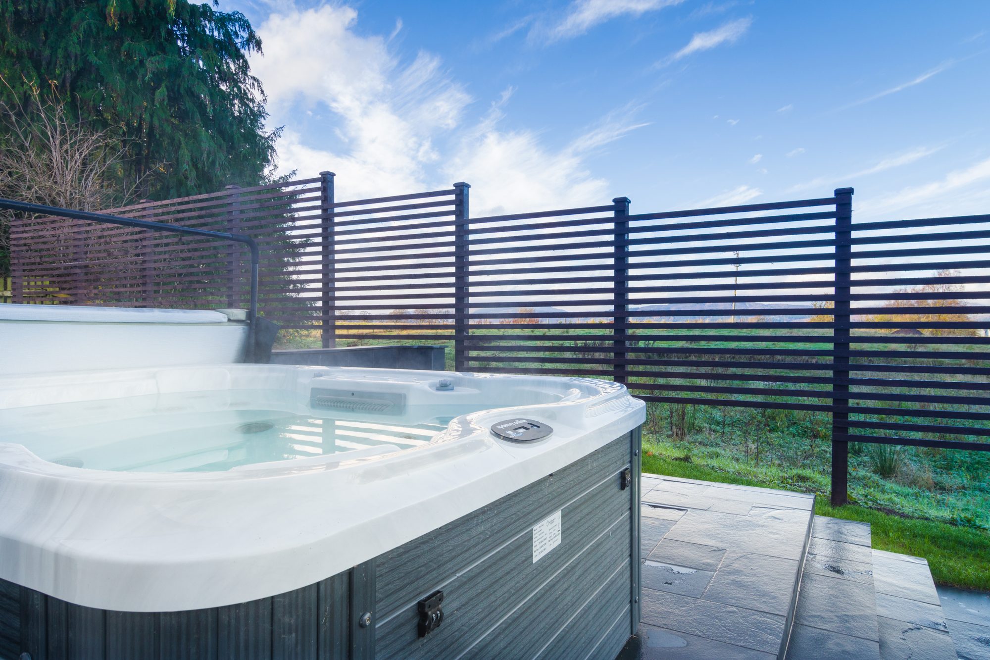 Outdoor hot tub on slate patio behind a dark slatted privacy fence with hills in the distance