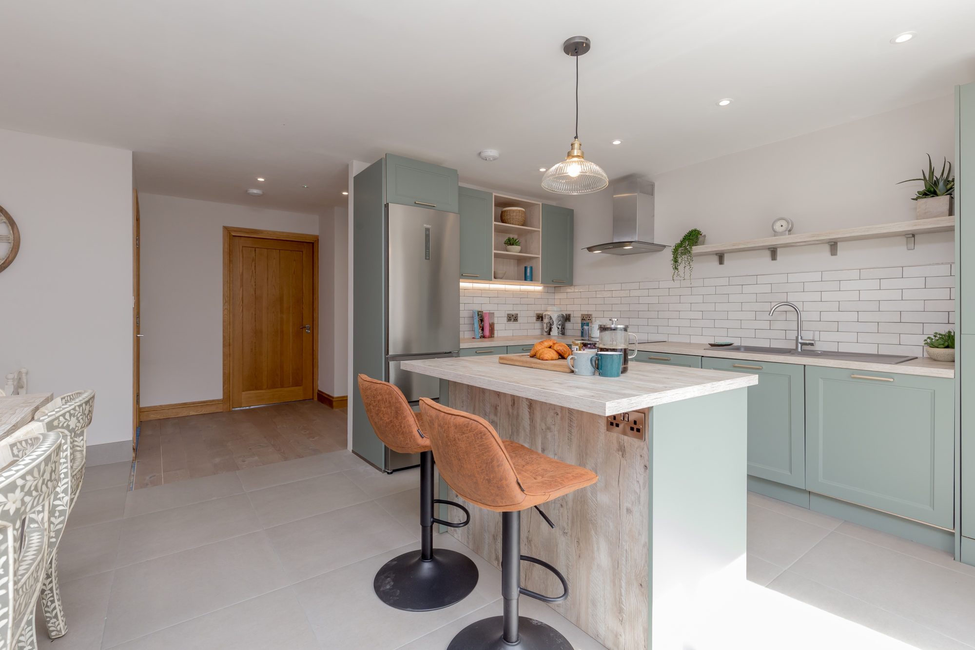 Sage shaker kitchen with pale wood island, tan leather bar stools and white metro tile splashback