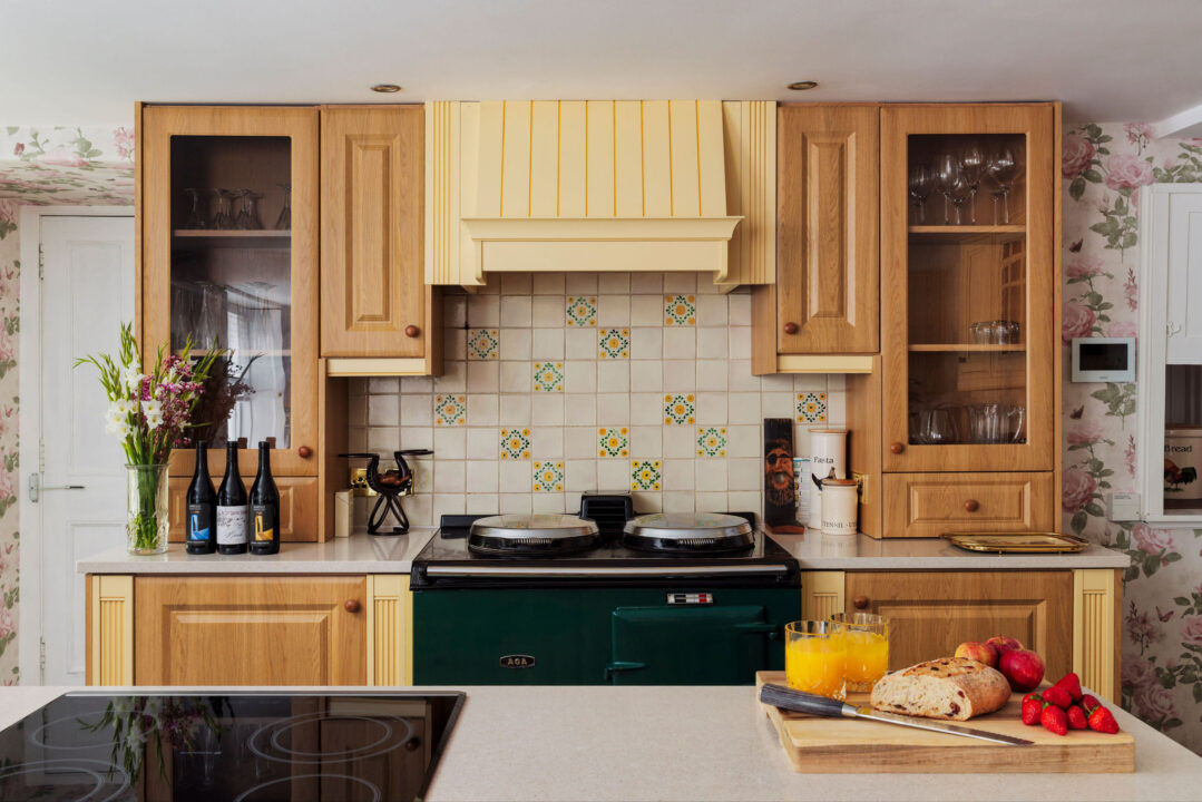 The Aga kitchen with oak cabinets, a green Aga, floral wallpaper and fresh bread, fruit and orange juice laid out on the island