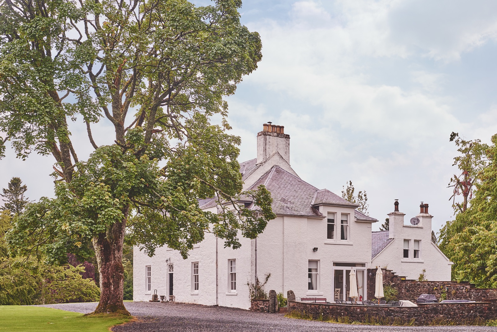 Side of the white lodge with its slate roof and chimneys, gravel drive and walled patio