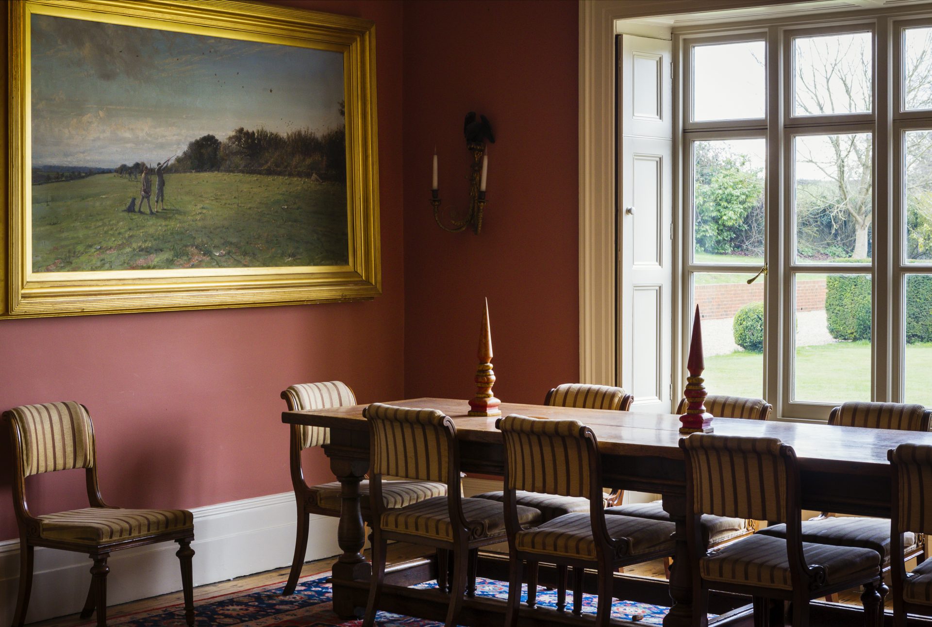 Country dining room with terracotta walls, gilt-framed pastoral painting and antique striped chairs, The Country Social