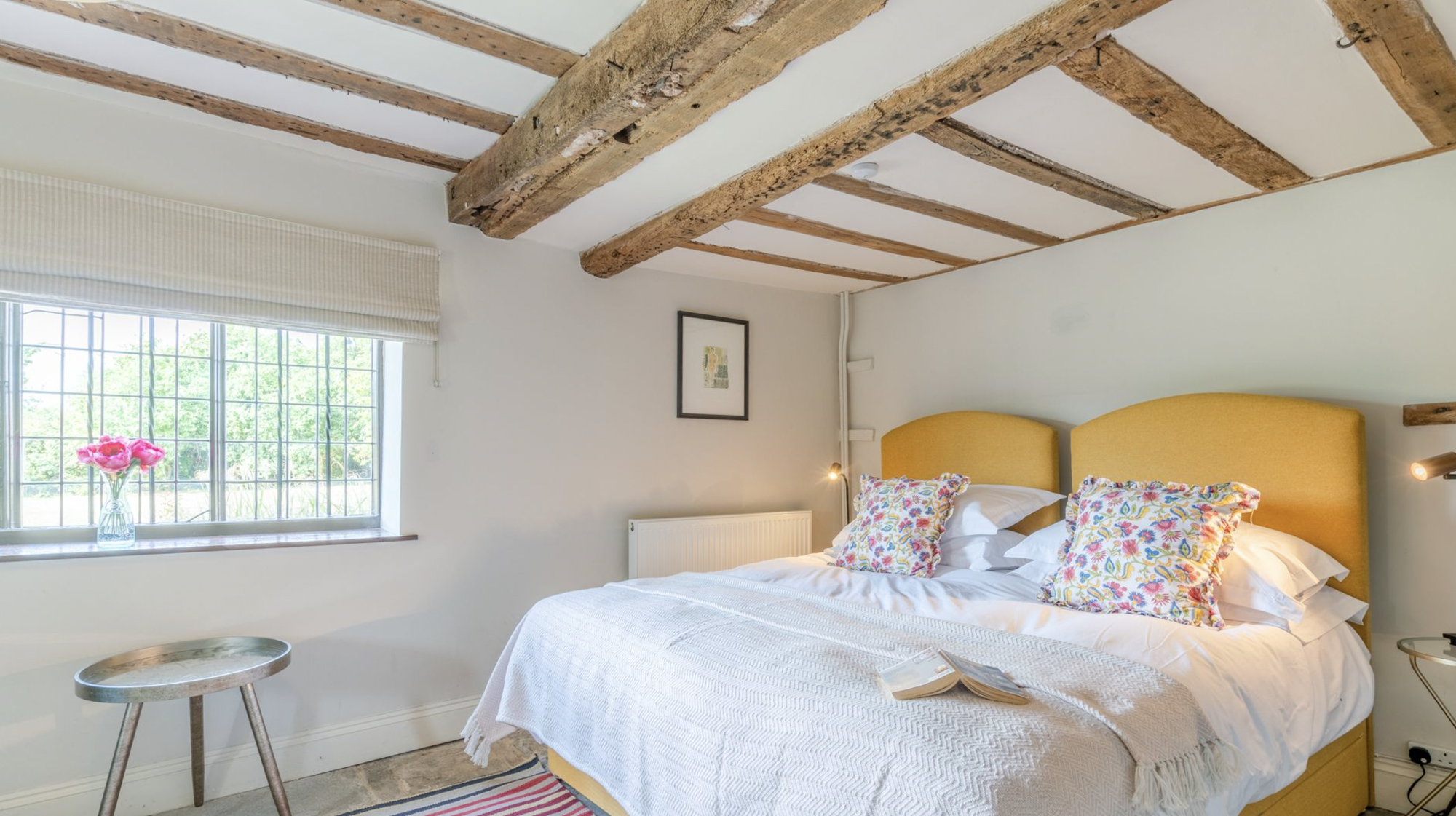 Cottage bedroom under thick oak ceiling beams with mustard headboard and leaded window