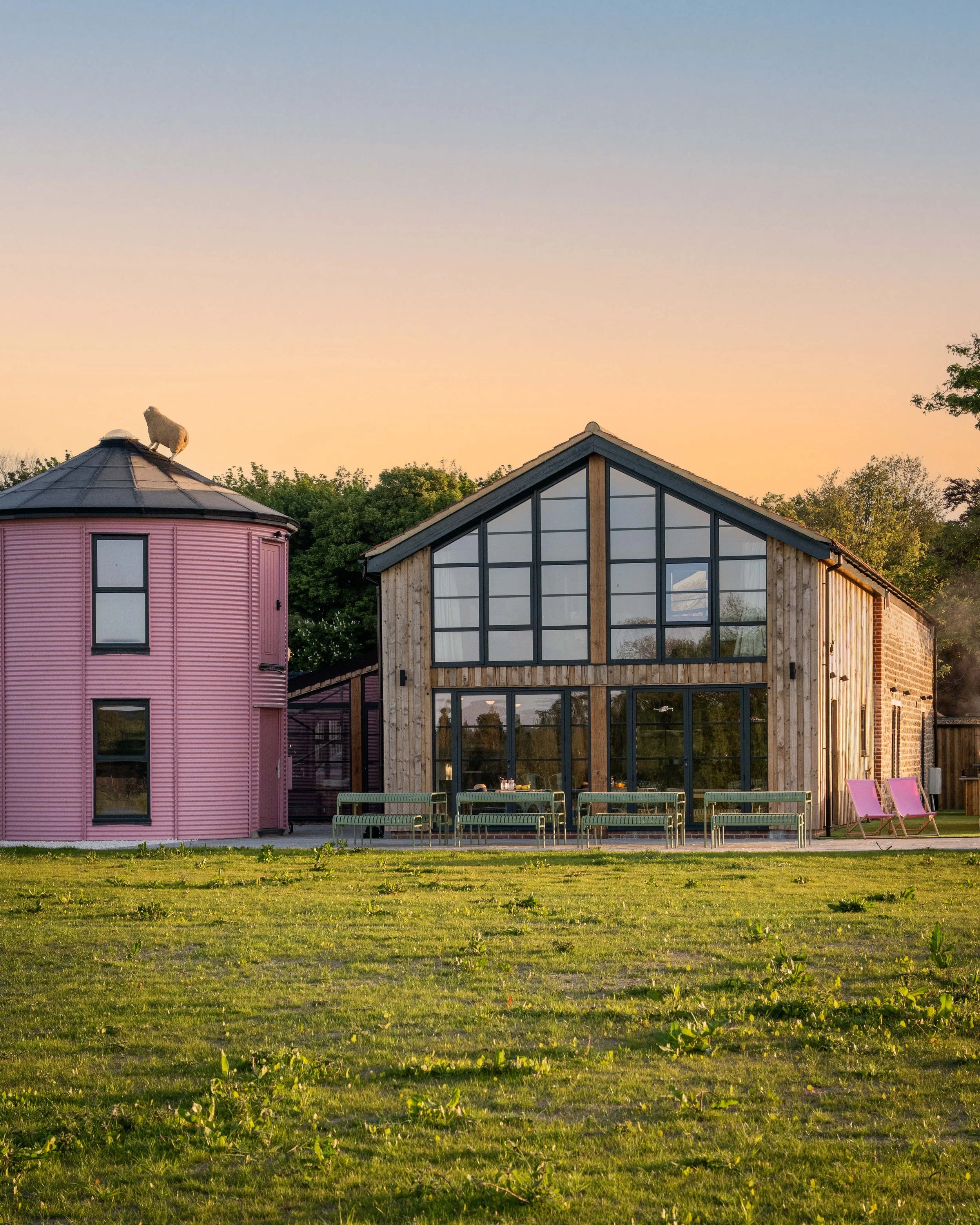 Pink circular tower beside the timber-clad gable barn at golden hour, with green outdoor benches lined up across the patio