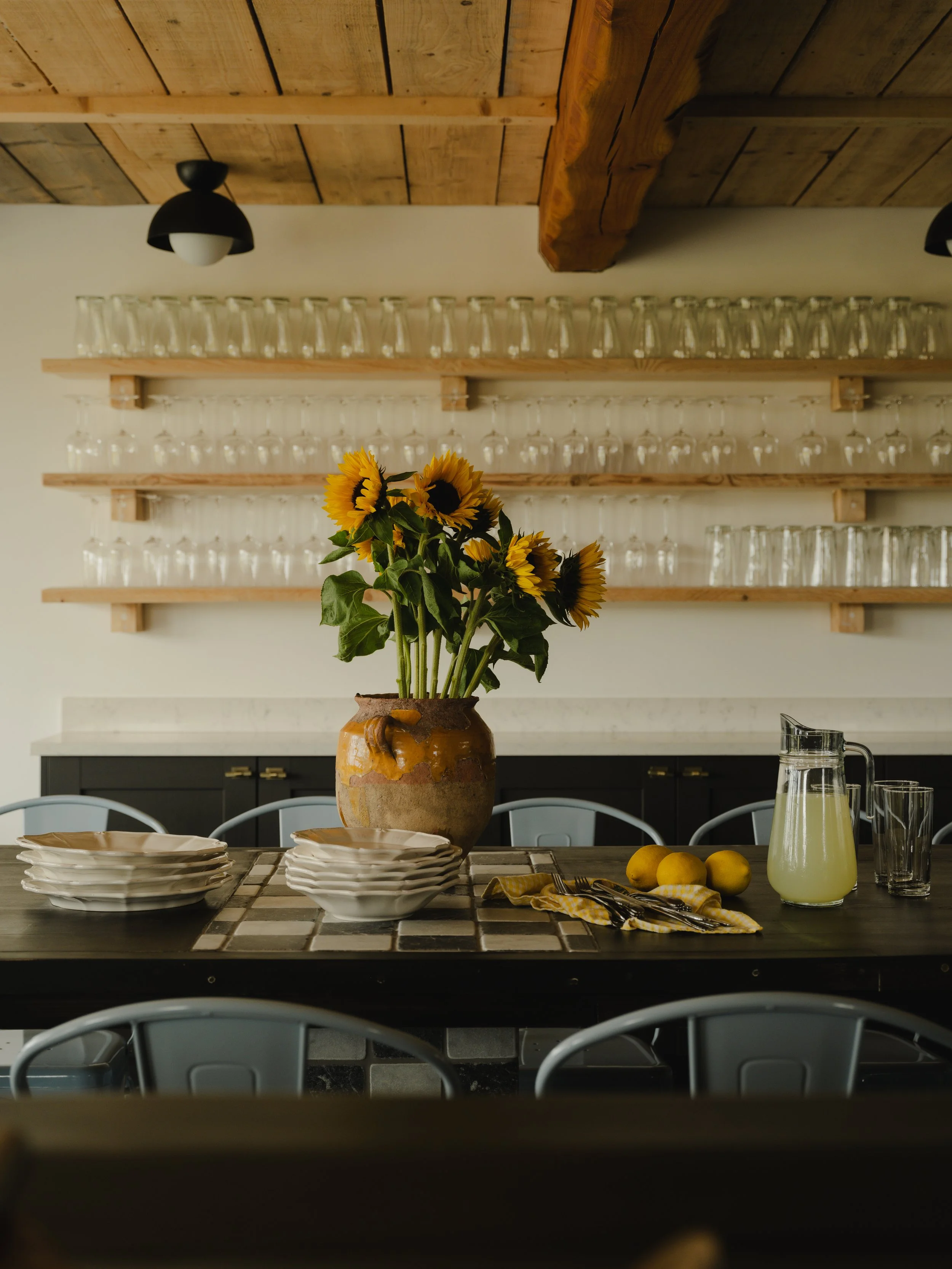 Long farmhouse table with sunflowers, stacked ceramic bowls, lemons and fresh lemonade under shelves of upturned wine glasses